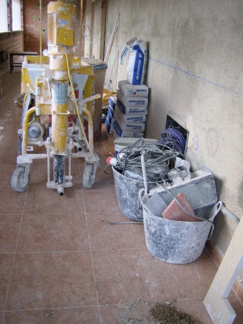 Máquina de proyectar yeso junto a sacos, cubos y cables en un pasillo.